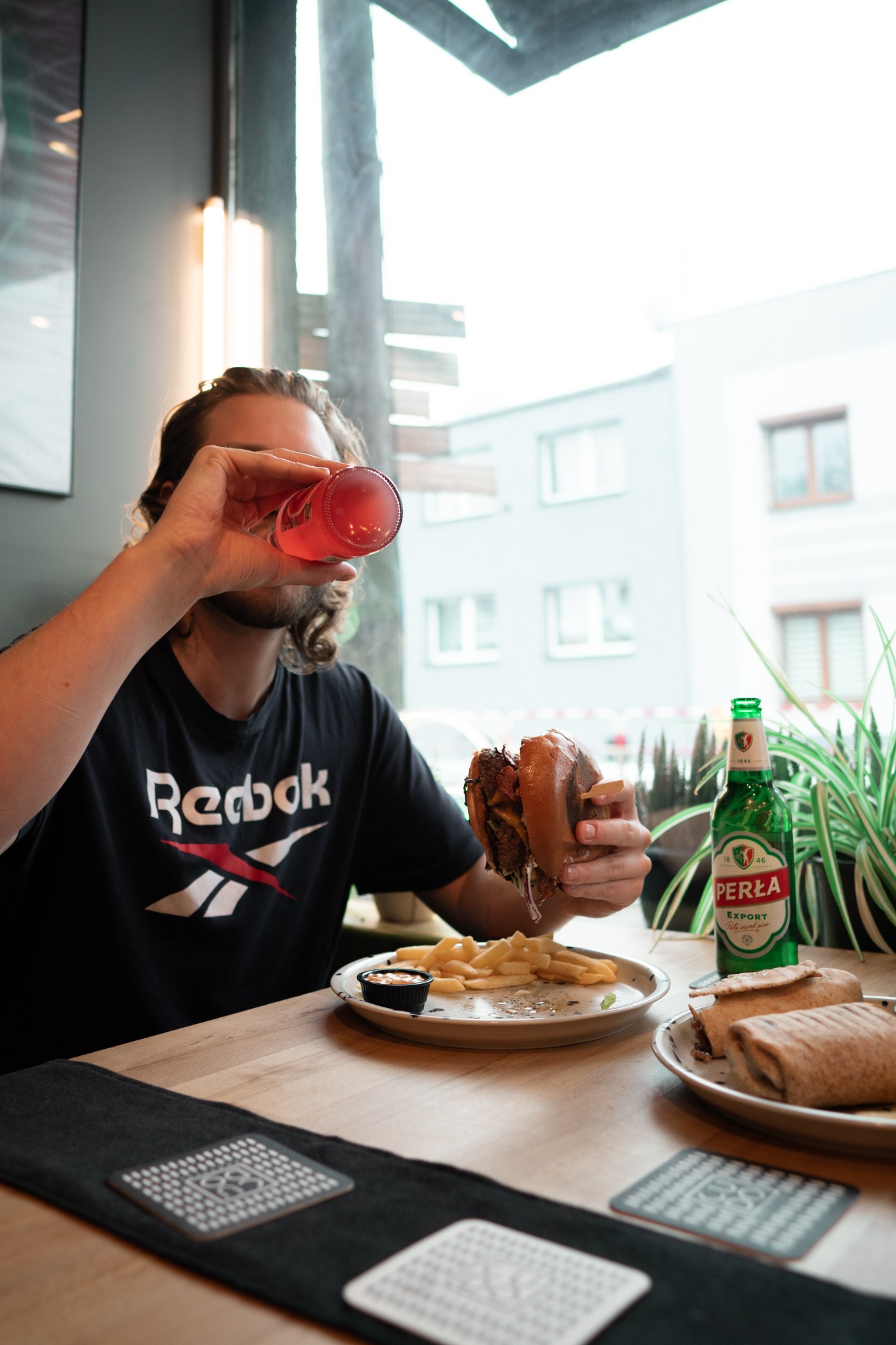 Mężczyzna w czarnej koszulce z logo Reebok pije różowy napój, trzymając burgera w ręku, na stole frytki, sos, tortilla i butelka piwa Perła Export.