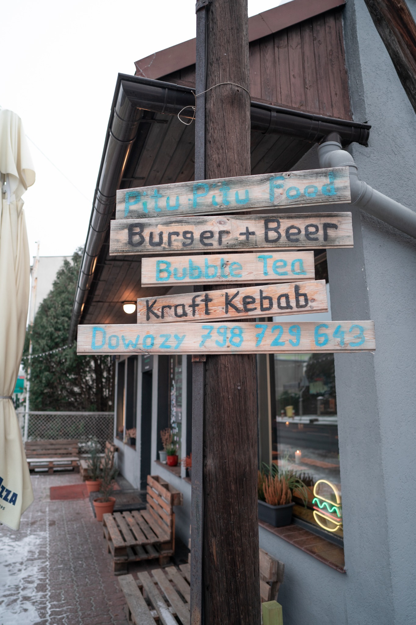 Drewniany słup z ręcznie malowanymi szyldami reklamującymi menu: Pitu Pitu Food, Burger + Beer, Bubble Tea, Kraft Kebab, Dowozy 798 729 643. W tle widoczny fragment budynku z neonem burgera w oknie.