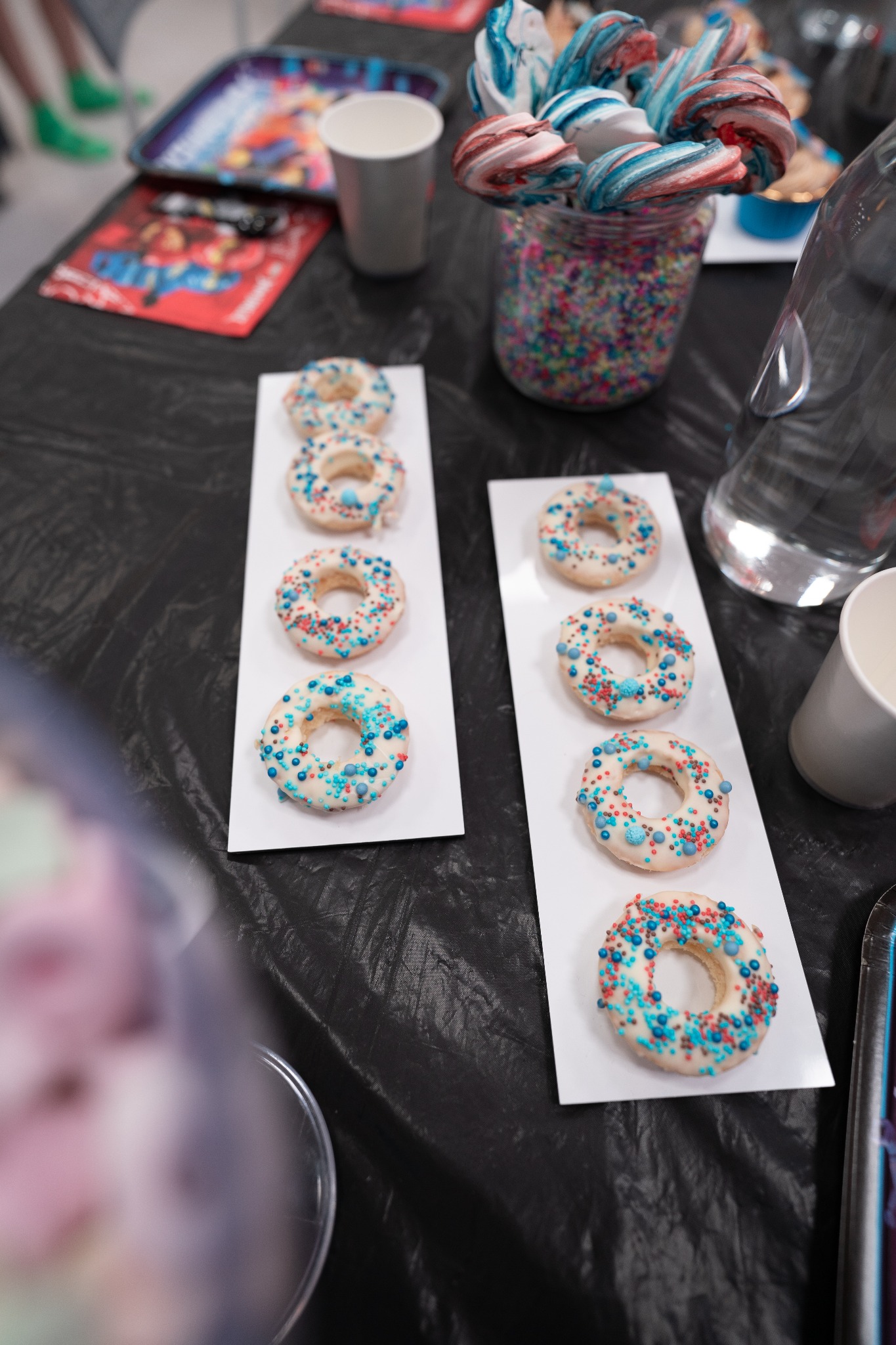 Dekorowane pączki na białych podkładkach, kolorowe lizaki w słoiku i papierowe kubki na czarnym, pomarszczonym tle stołu.
