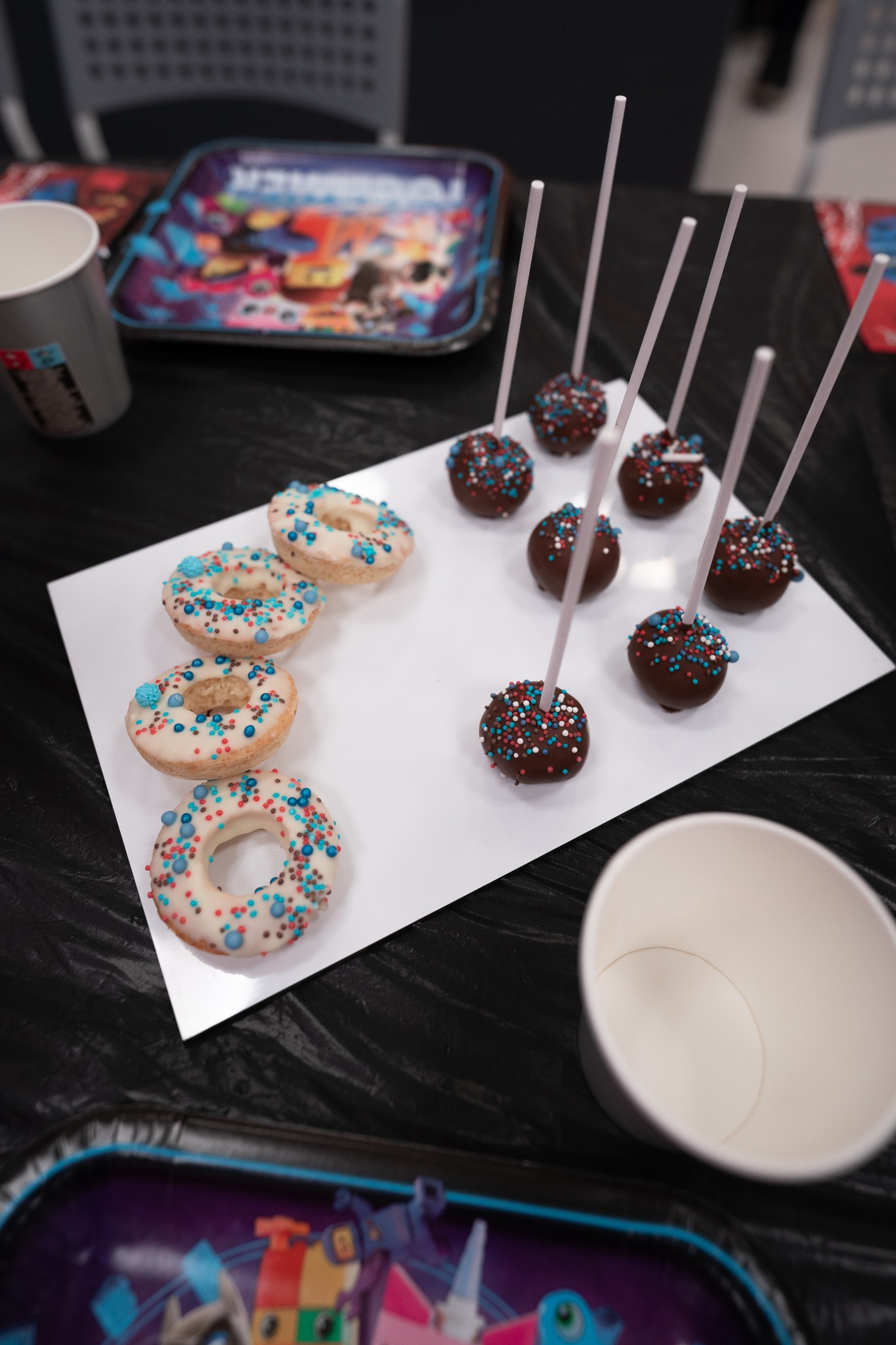 Donuty z białym lukrem i kolorową posypką oraz czekoladowe cake pops z patyczkami na białej tacy, obok kubki i tacki z motywami bajkowymi, na czarnym stole.