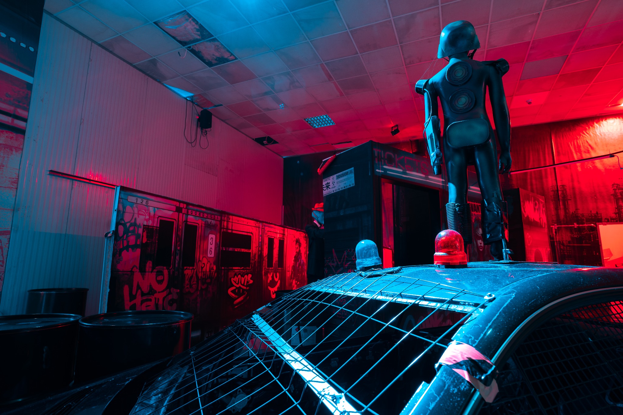 Scena z escape roomu: robot w futurystycznym stroju stoi na dachu z kratą, czerwona lampa ostrzegawcza, graffiti na wagonie metra w tle, oświetlenie neonowe w odcieniach czerwieni i błękitu.