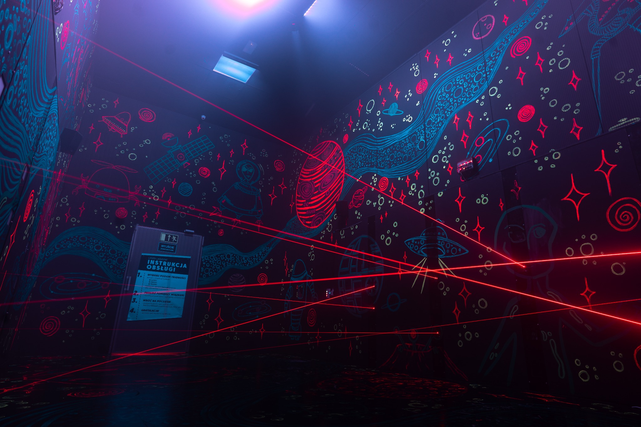 Wnętrze laserowego labiryntu z neonowymi rysunkami kosmosu na ścianach, oświetlone czerwonymi wiązkami lasera i instrukcją obsługi.