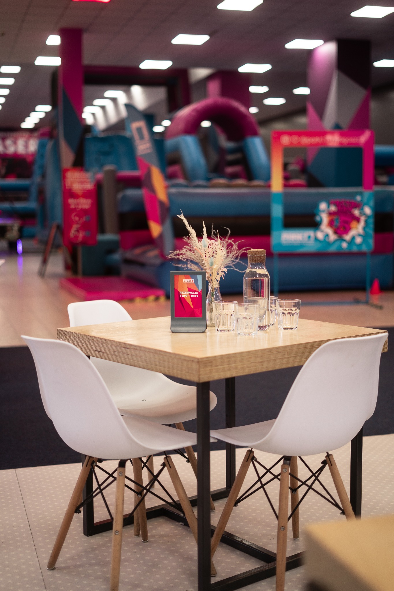Stół z czterema białymi krzesłami w tle sali zabaw z dmuchanymi konstrukcjami, na stole wazonik z suszem, karafka z wodą, szklanki i tablet z rezerwacją.