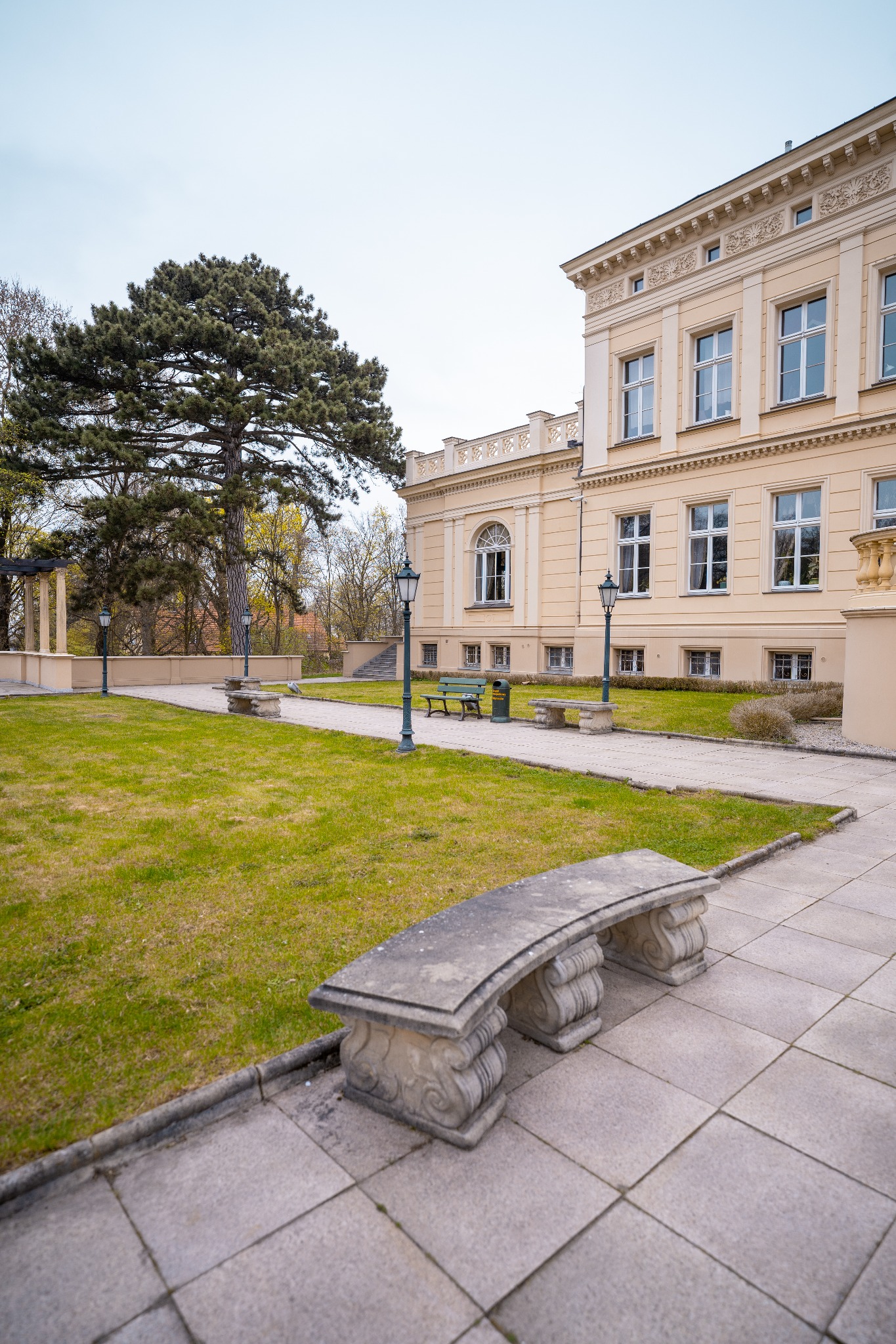 Klasycystyczny pałac z kremową fasadą, park z ławkami z piaskowca i zielenią, oświetlenie parkowe w stylu retro.