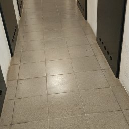 Perfekcyjne Sprzątanie MAŁGORZATA WRÓBEL - Długi korytarz z szarymi płytkami podłogowymi i rzędem czarnych drzwi z otworami wentylacyjnymi.