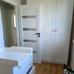 Perfekcyjne Sprzątanie MAŁGORZATA WRÓBEL - Pokój dziecięcy z białymi meblami: komoda z otwartą szufladą i trzydrzwiowa szafa z jasnymi uchwytami, widoczny kosz na śmieci w rogu.