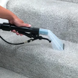 Czyszczenie dywanu na schodach za pomocą profesjonalnego ekstraktora z przezroczystą dyszą natryskową, widoczna ręka operatora.