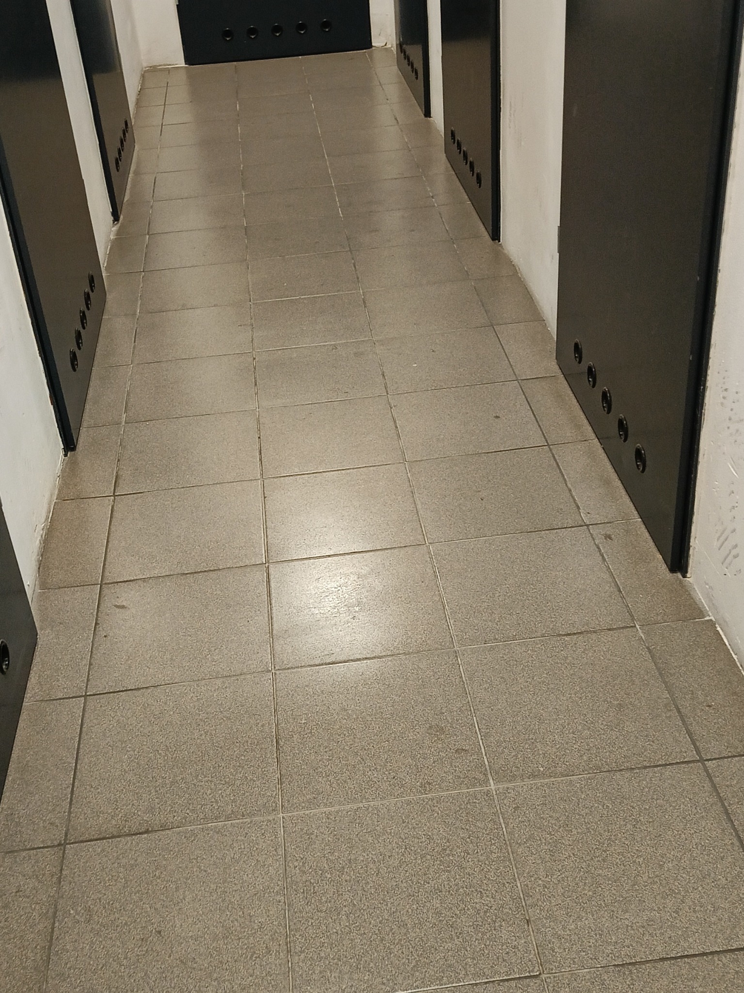 Długi korytarz z szarymi płytkami podłogowymi i rzędem czarnych drzwi z otworami wentylacyjnymi.