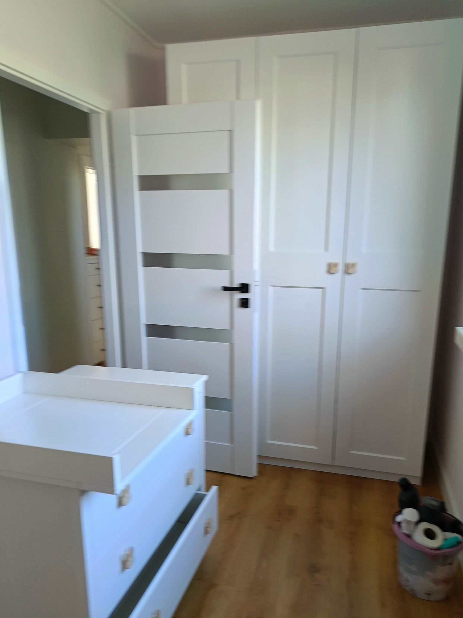 Pokój dziecięcy z białymi meblami: komoda z otwartą szufladą i trzydrzwiowa szafa z jasnymi uchwytami, widoczny kosz na śmieci w rogu.