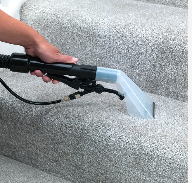 Czyszczenie dywanu na schodach za pomocą profesjonalnego ekstraktora z przezroczystą dyszą natryskową, widoczna ręka operatora.