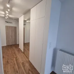 Biała zabudowa wnękowa z lustrami w przedpokoju z drewnianą podłogą, widoczny grzejnik i logo firmy meblarskiej w prawym dolnym rogu.