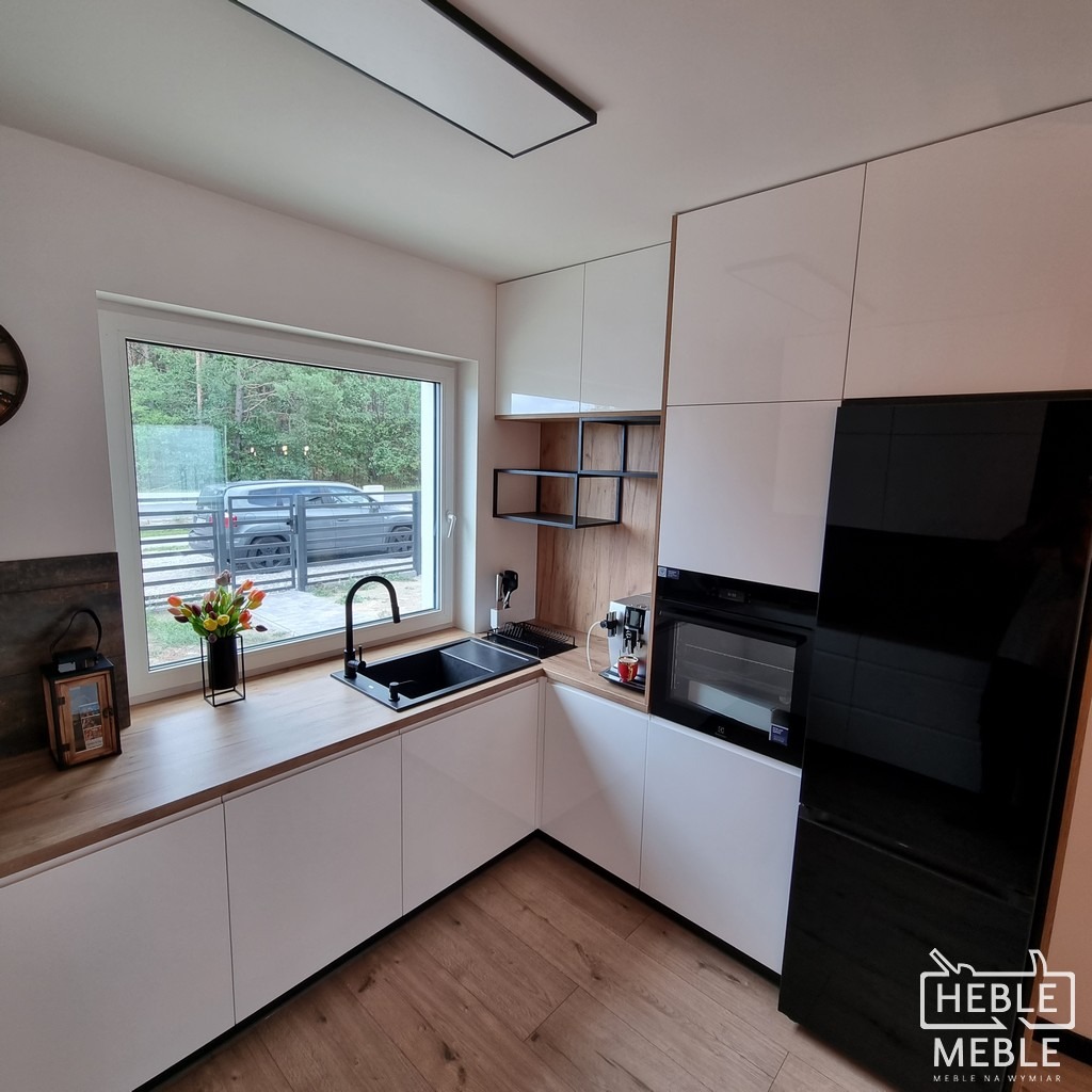 Nowoczesna kuchnia z białymi, lakierowanymi frontami, drewnianym blatem i czarnym zlewozmywakiem pod oknem z widokiem na zieleń; obok zabudowana czarna lodówka i piekarnik.