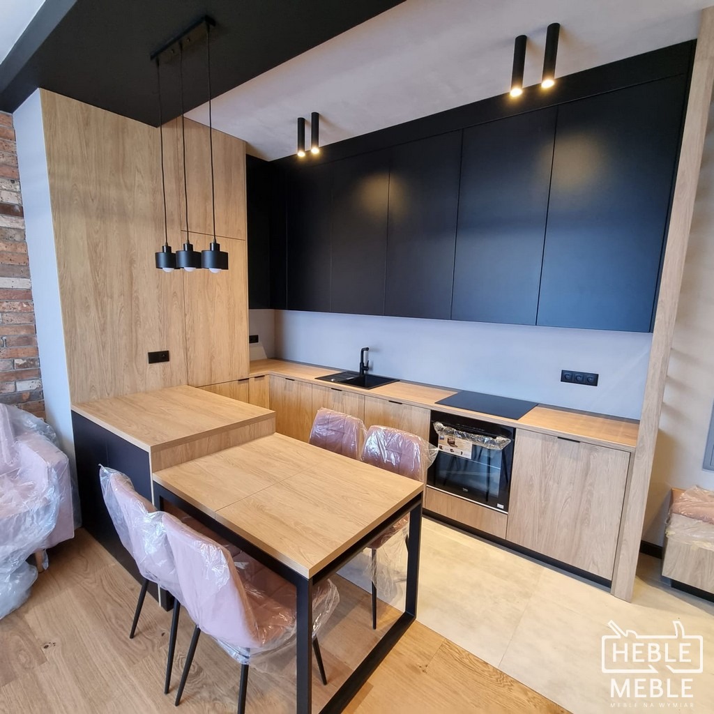 Nowoczesna kuchnia z czarnymi górnymi szafkami i drewnopodobnymi dolnymi, połączona z jadalnią ze stołem i krzesłami w folii ochronnej, oświetlona wiszącymi lampami.