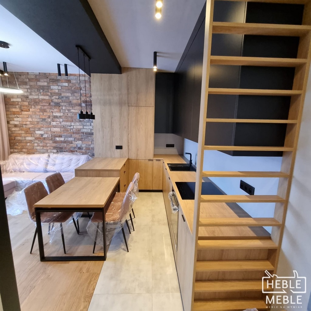 Nowoczesna kuchnia z drewnianymi frontami i czarnymi akcentami, widok na stół jadalny i ceglaną ścianę w tle, krzesła zabezpieczone folią ochronną, drewniana drabina prowadząca do antresoli po...