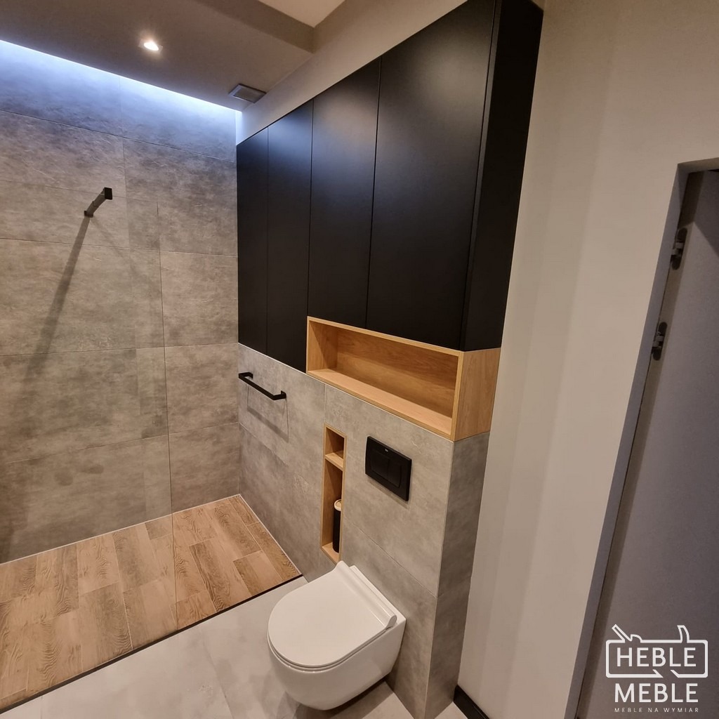 Nowoczesna łazienka z czarnymi szafkami wiszącymi nad toaletą, drewnianą półką i szarymi płytkami, widoczny fragment kabiny prysznicowej z drewnopodobną podłogą i logo firmy w prawym dolnym rogu.