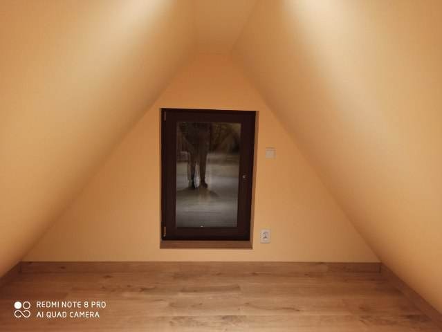 Poddasze ze skośnymi ścianami pomalowanymi na kremowo, drewniana podłoga, okno z ciemną ramą, widoczna instalacja elektryczna.