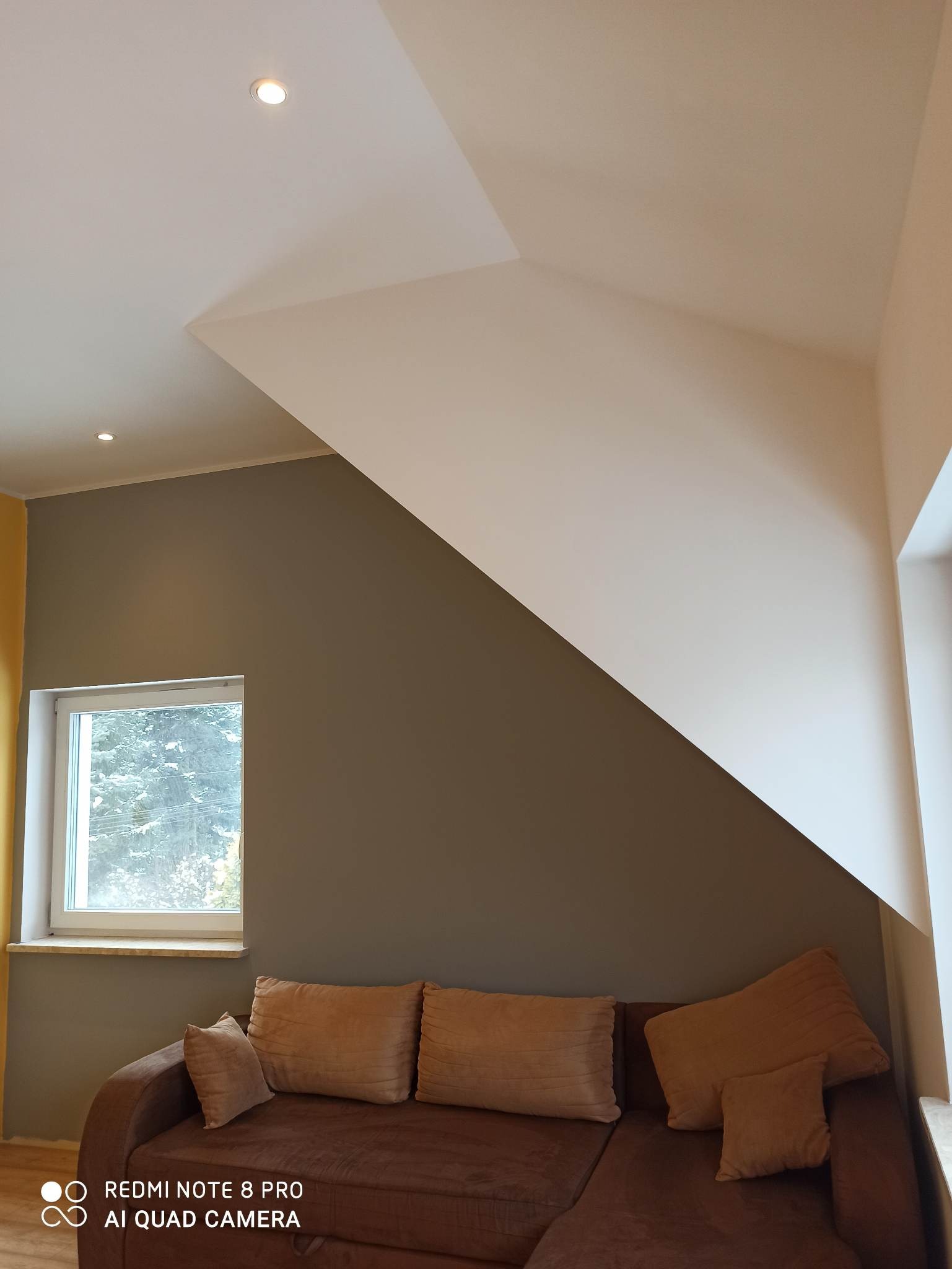 Pokój na poddaszu po malowaniu, z brązową kanapą, oknem i skośnym sufitem z oświetleniem punktowym.