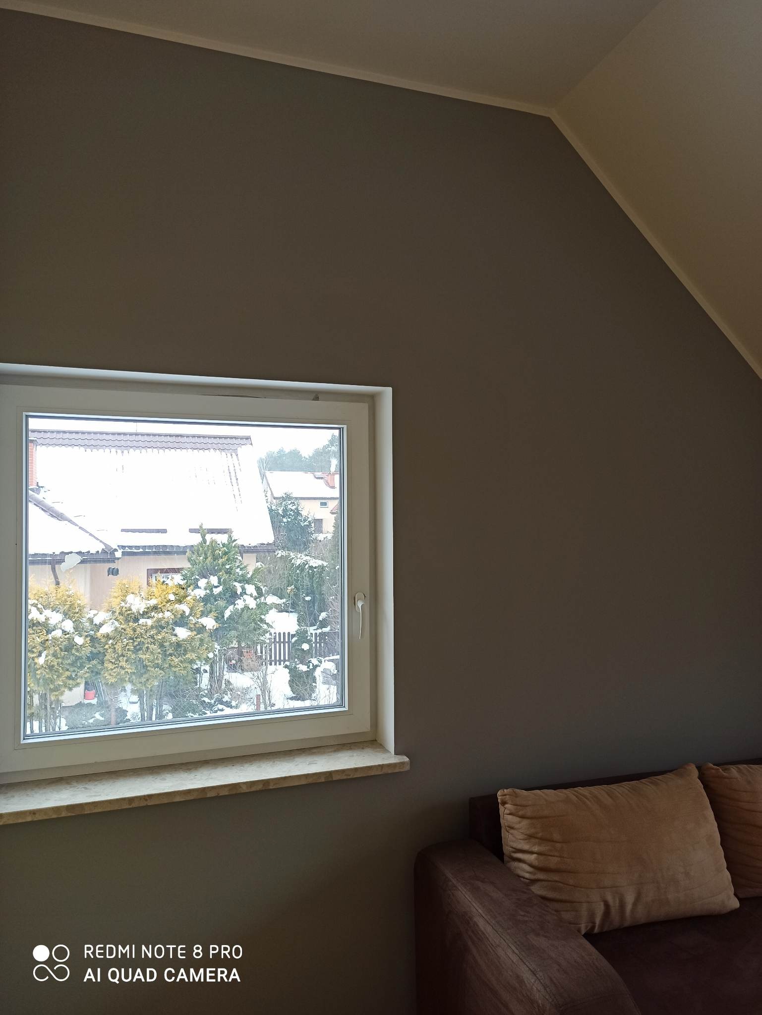 Szaro-brązowa ściana w pokoju po malowaniu, widoczne okno z widokiem na ośnieżony ogród i fragment sofy z poduszkami.