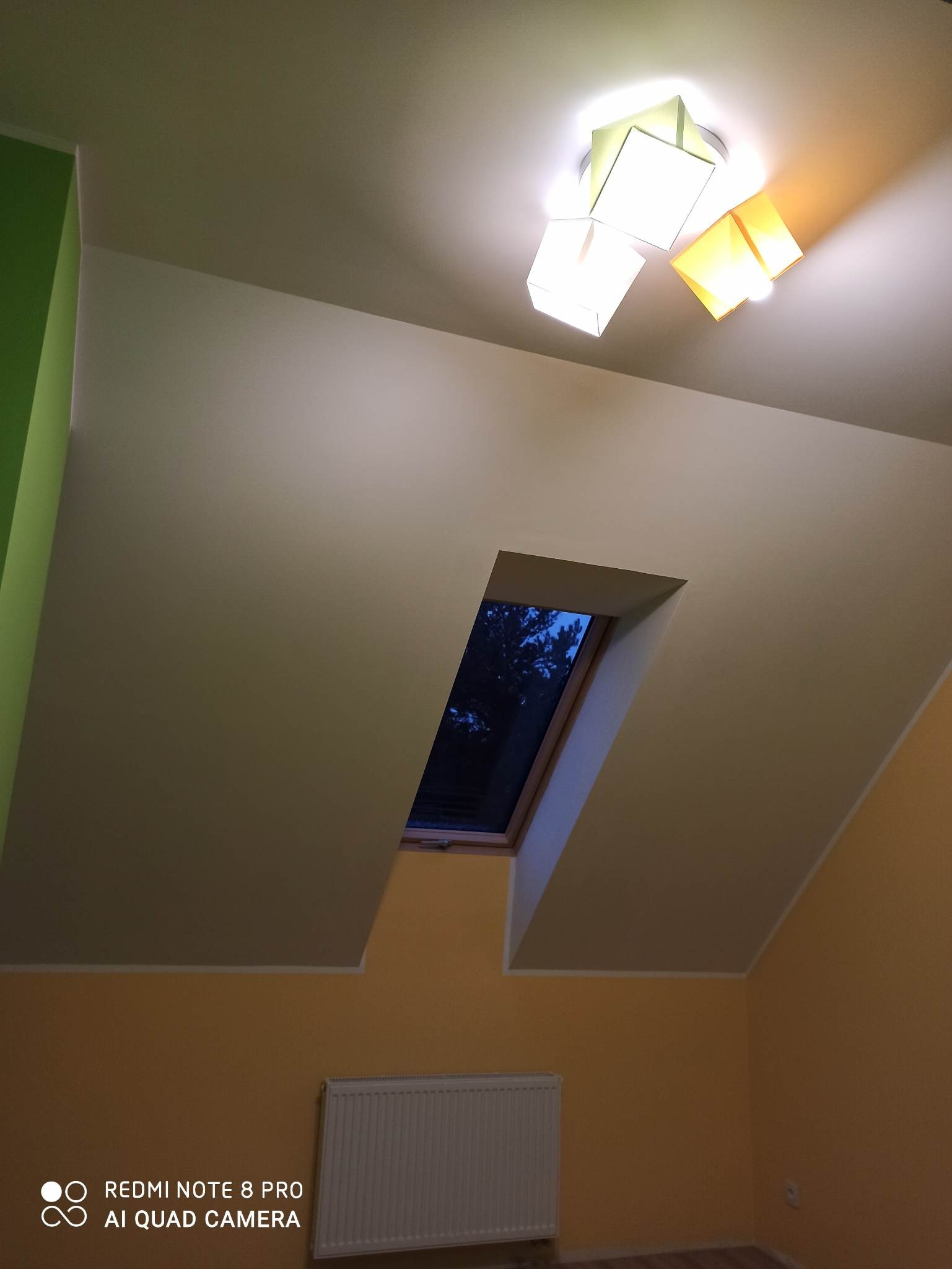 Poddasze z oknem dachowym, grzejnikiem i nowoczesną lampą sufitową z kwadratowymi kloszami w kolorach białym, zielonym i żółtym. Ściany pomalowane na żółto i kremowo, z zielonym akcentem na jednej...