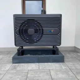 Czarna pompa ciepła na betonowej podstawie, zainstalowana na zewnątrz budynku z szarą kostką brukową, widoczny fragment brązowych drzwi i okna.