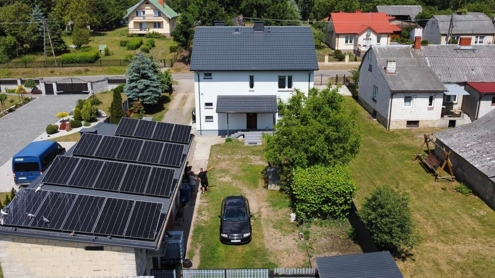 Widok z drona na dom z zainstalowanymi panelami słonecznymi na dachu garażu, obok stoi czarny samochód, a w tle widać dwóch mężczyzn.