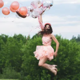 Uśmiechnięta kobieta w różowej sukience i wianku na głowie skacze na trawie, trzymając w ręku balony w odcieniach różu i przezroczyste z konfetti, w tle zielone drzewa i błękitne niebo.