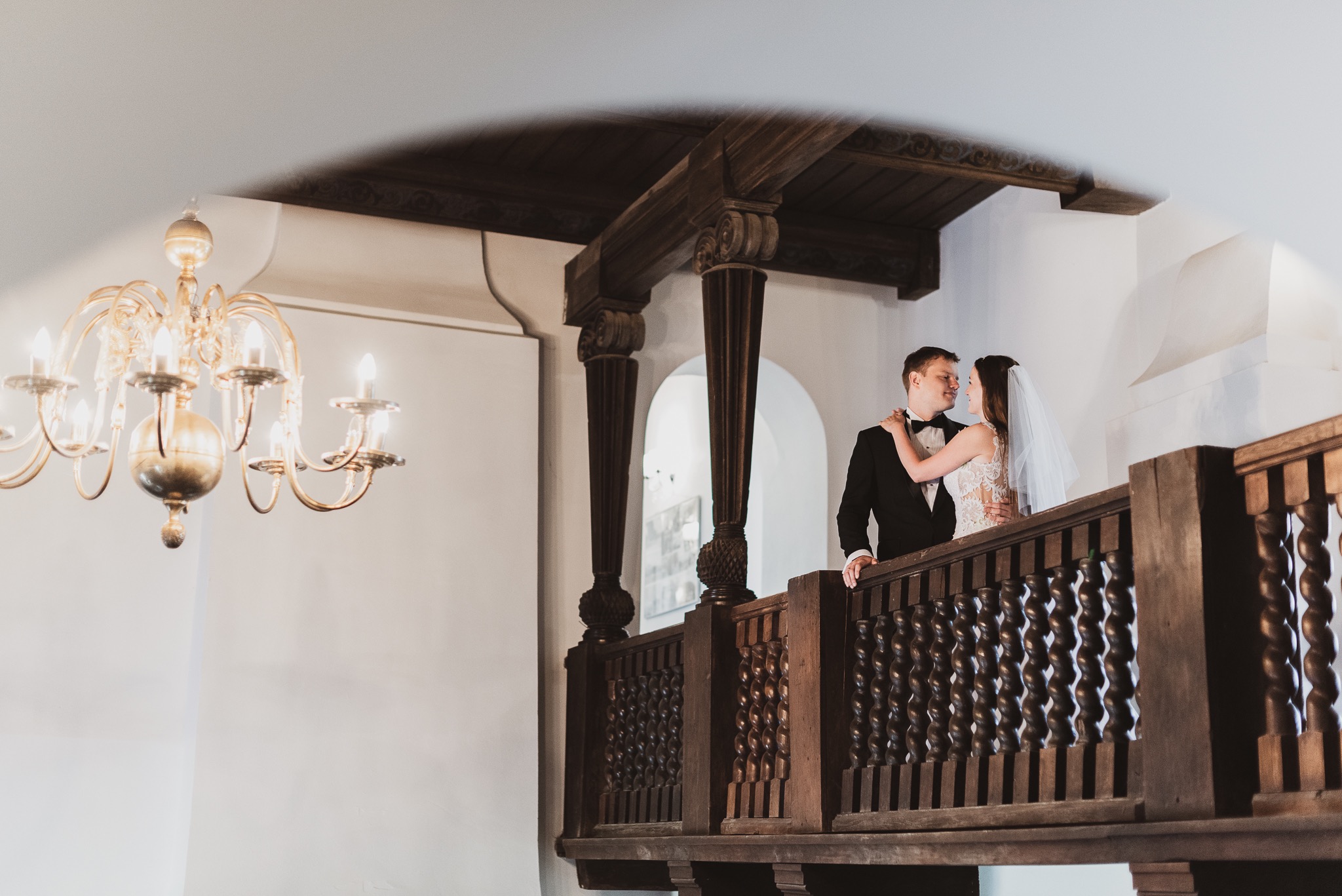 Para młoda na drewnianym balkonie z ozdobną balustradą, on w czarnym smokingu, ona w koronkowej sukni ślubnej i welonie, w tle białe ściany i złoty żyrandol.