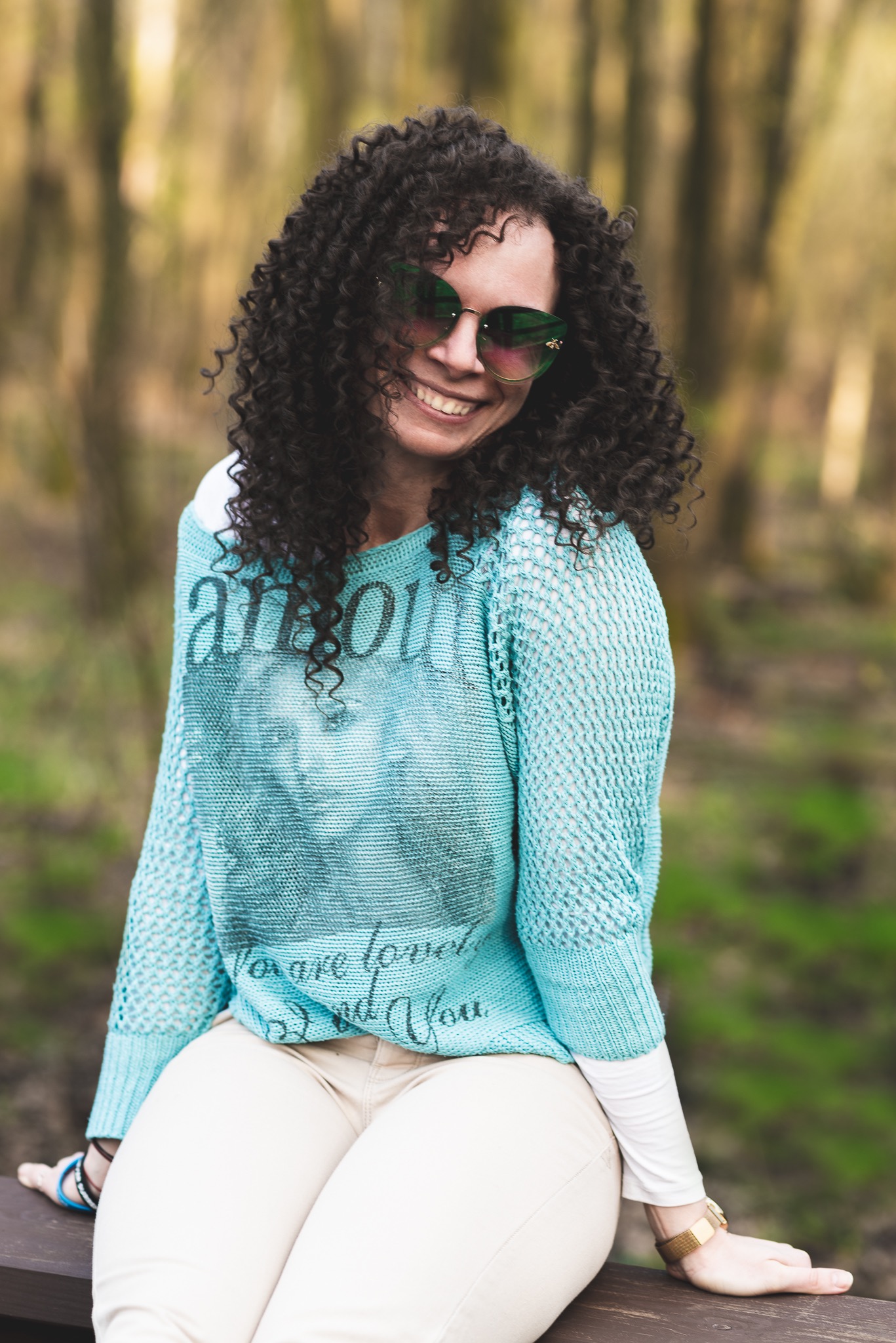 Uśmiechnięta kobieta z kręconymi włosami i okularami przeciwsłonecznymi siedzi na drewnianej ławce w lesie, ubrana w ażurowy, turkusowy sweter z napisem.