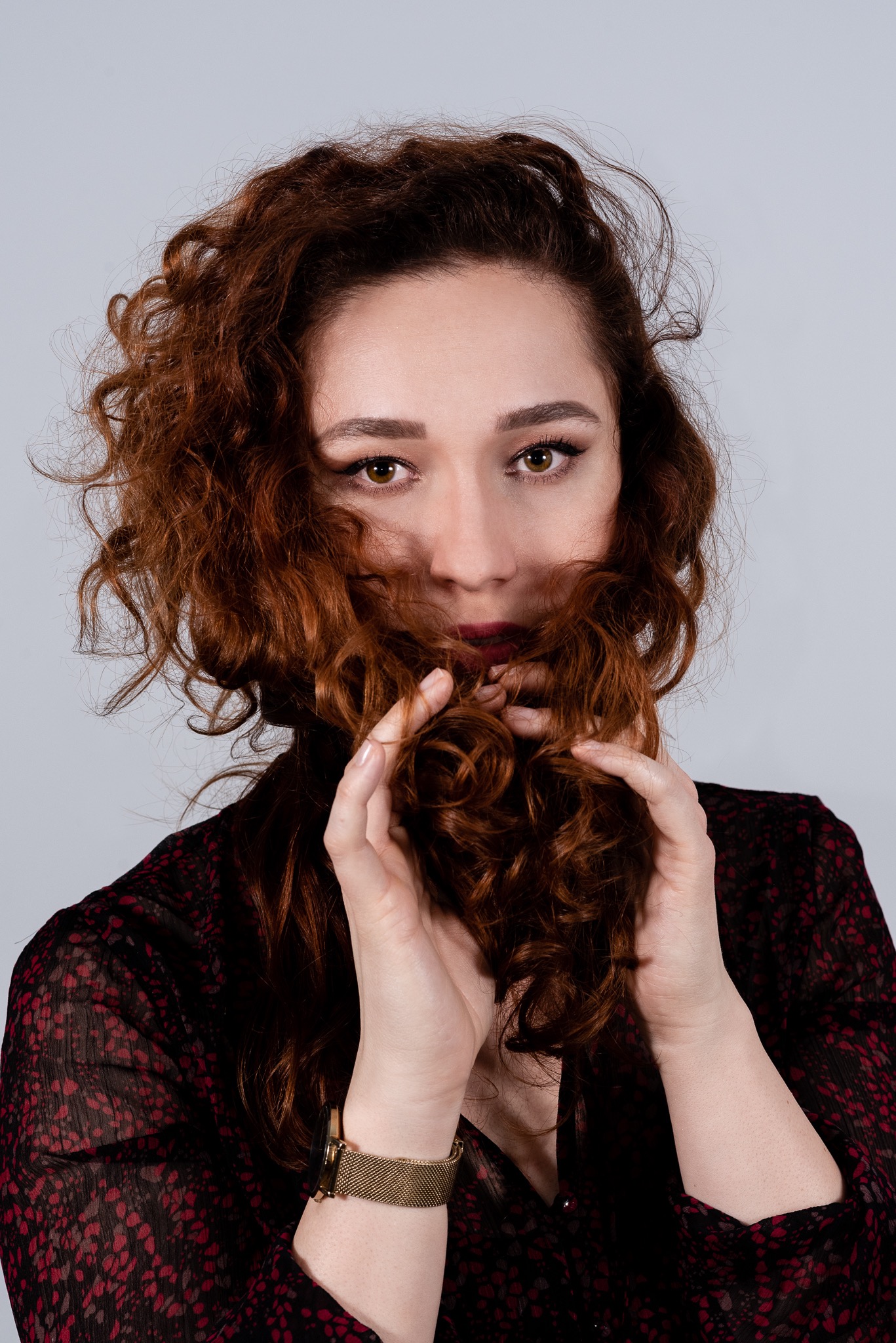Portret kobiety z rudymi kręconymi włosami, częściowo zakrywającymi twarz, w ciemnej bluzce w Zbójnej, z delikatnym makijażem i złotym zegarkiem na nadgarstku.