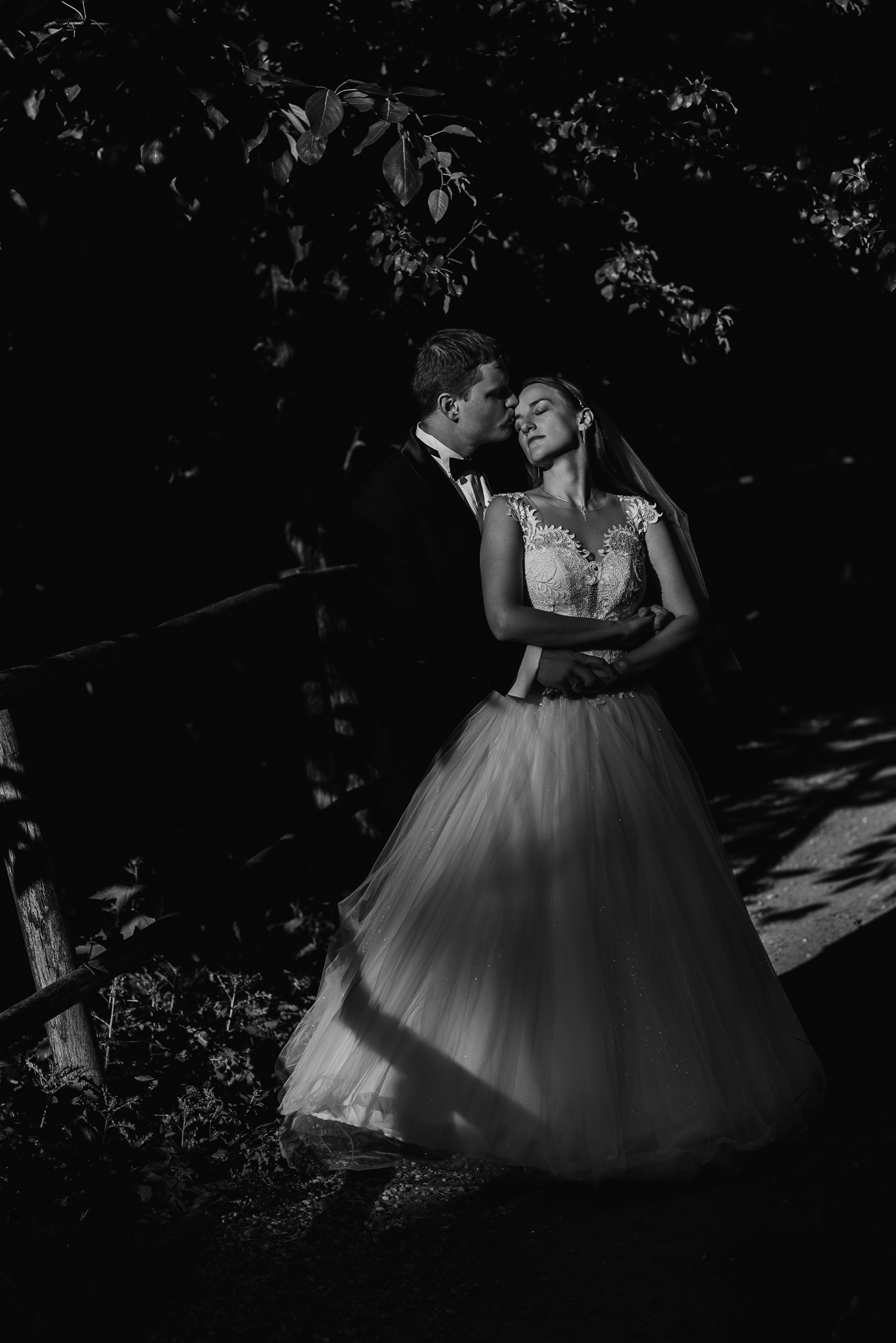 Czarno-białe zdjęcie pary młodej: pan młody w smokingu całuje pannę młodą w koronkową suknię ślubną, oboje stoją w zacienionym plenerze z drewnianym płotkiem.