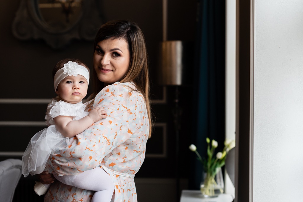 Uśmiechnięta kobieta w kwiecistej sukience trzymająca na rękach niemowlę w białej sukience i opasce z kwiatem, w tle wazon z białymi tulipanami.