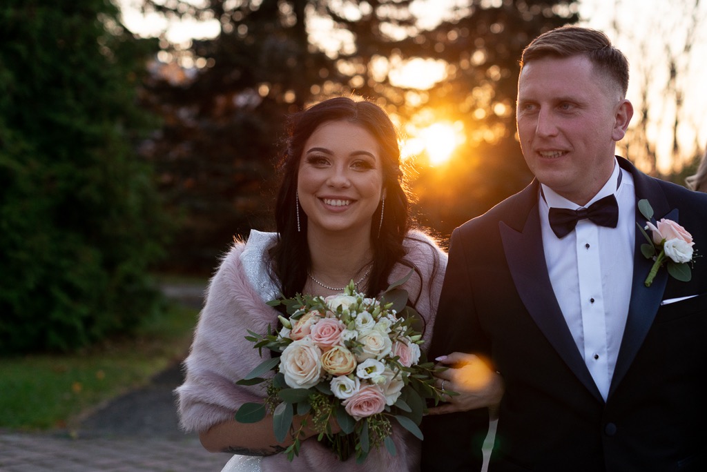 Uśmiechnięta panna młoda w różowym futerku, trzymająca bukiet z różowych i białych róż, idzie pod rękę z panem młodym w czarnym smokingu na tle zachodzącego słońca.