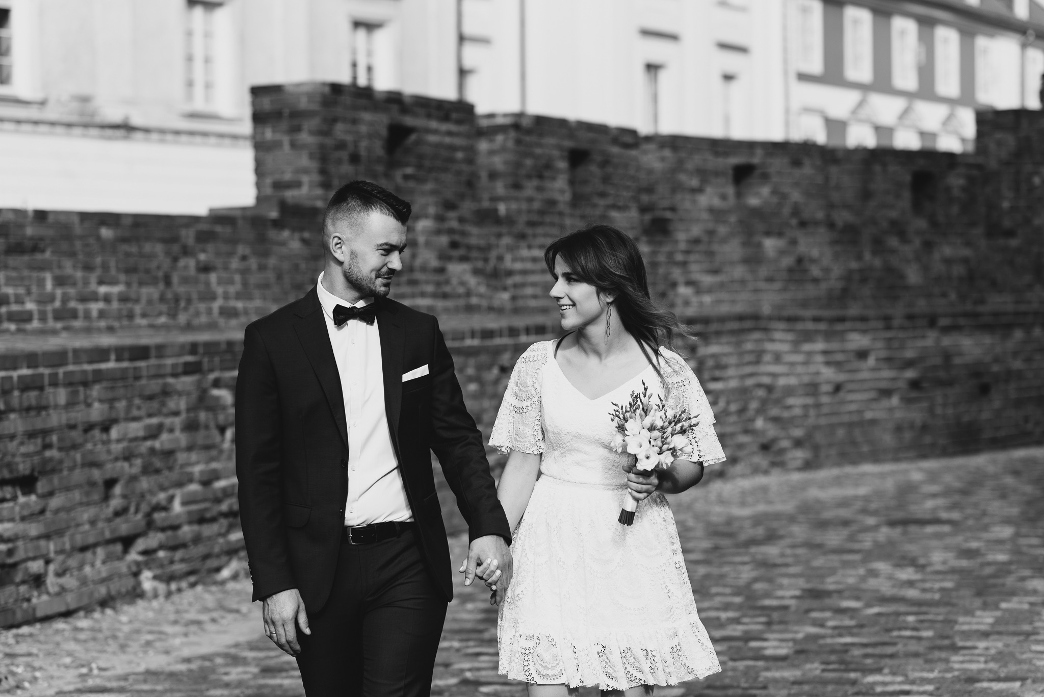 Czarno-białe zdjęcie uśmiechniętej pary młodej trzymającej się za ręce, idącej po brukowanej ulicy na tle ceglanego muru i budynków w oddali.