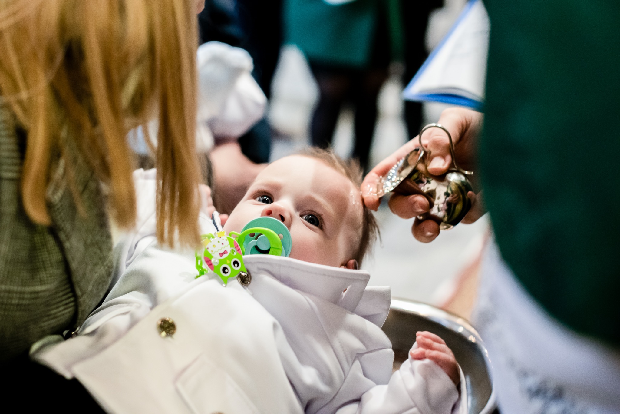 Niemowlę z zielonym smoczkiem, ubrane w białe ubranko, leży w metalowej misce, podczas gdy ksiądz kropi jego głowę wodą święconą.