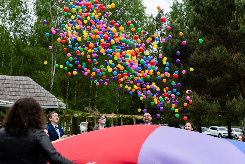 Grupa osób trzymająca kolorową płachtę, z której w górę wylatują setki wielobarwnych piłeczek na tle zieleni drzew.