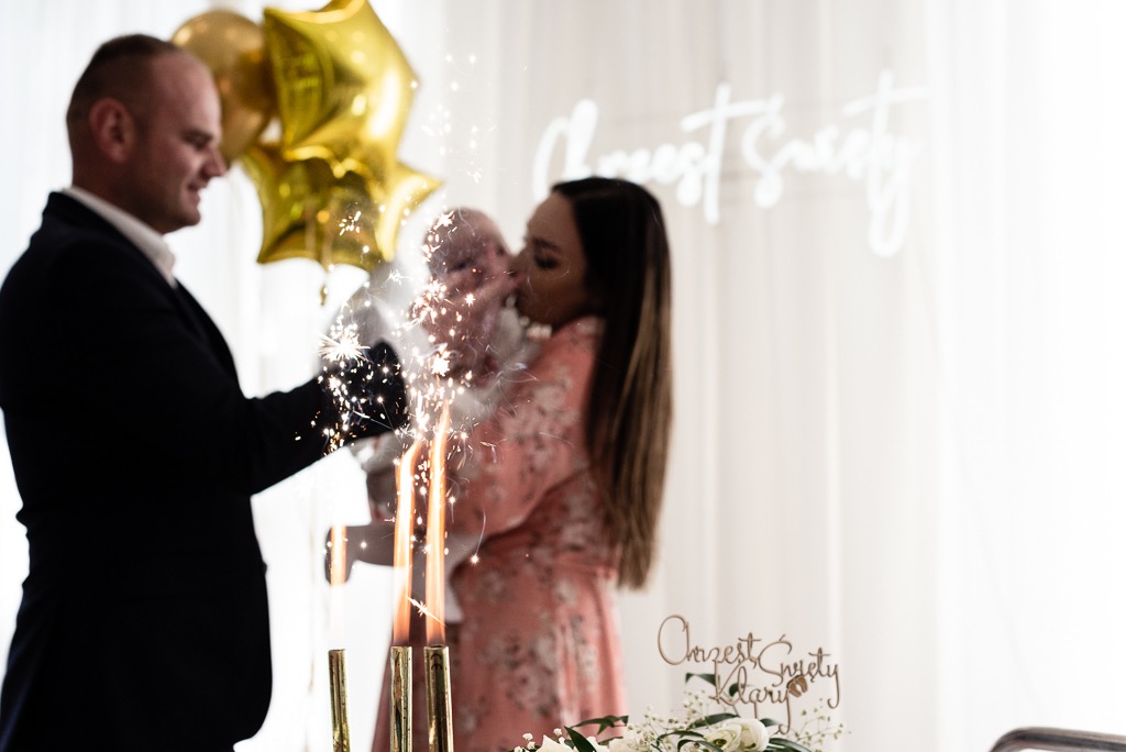 Rodzice świętują chrzest dziecka z fontannami tortowymi, złotymi balonami w kształcie gwiazd i napisem 'Chrzest Święty Klary' w tle.