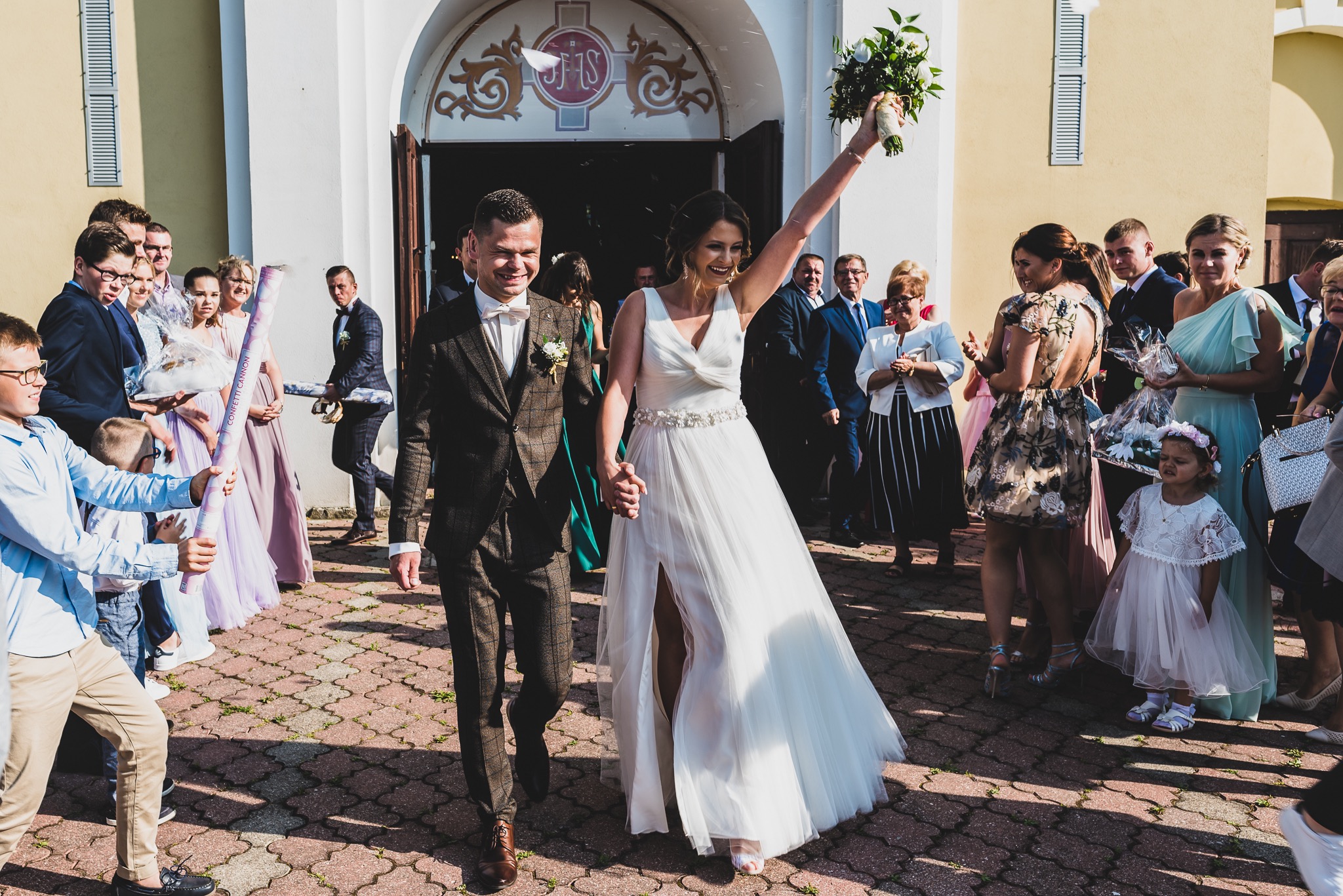 Uśmiechnięta para młoda wychodzi z kościoła, trzymając się za ręce, w towarzystwie gości z konfetti i kwiatami. Panna młoda w białej sukni z rozcięciem, pan młody w brązowym garniturze w kratę.