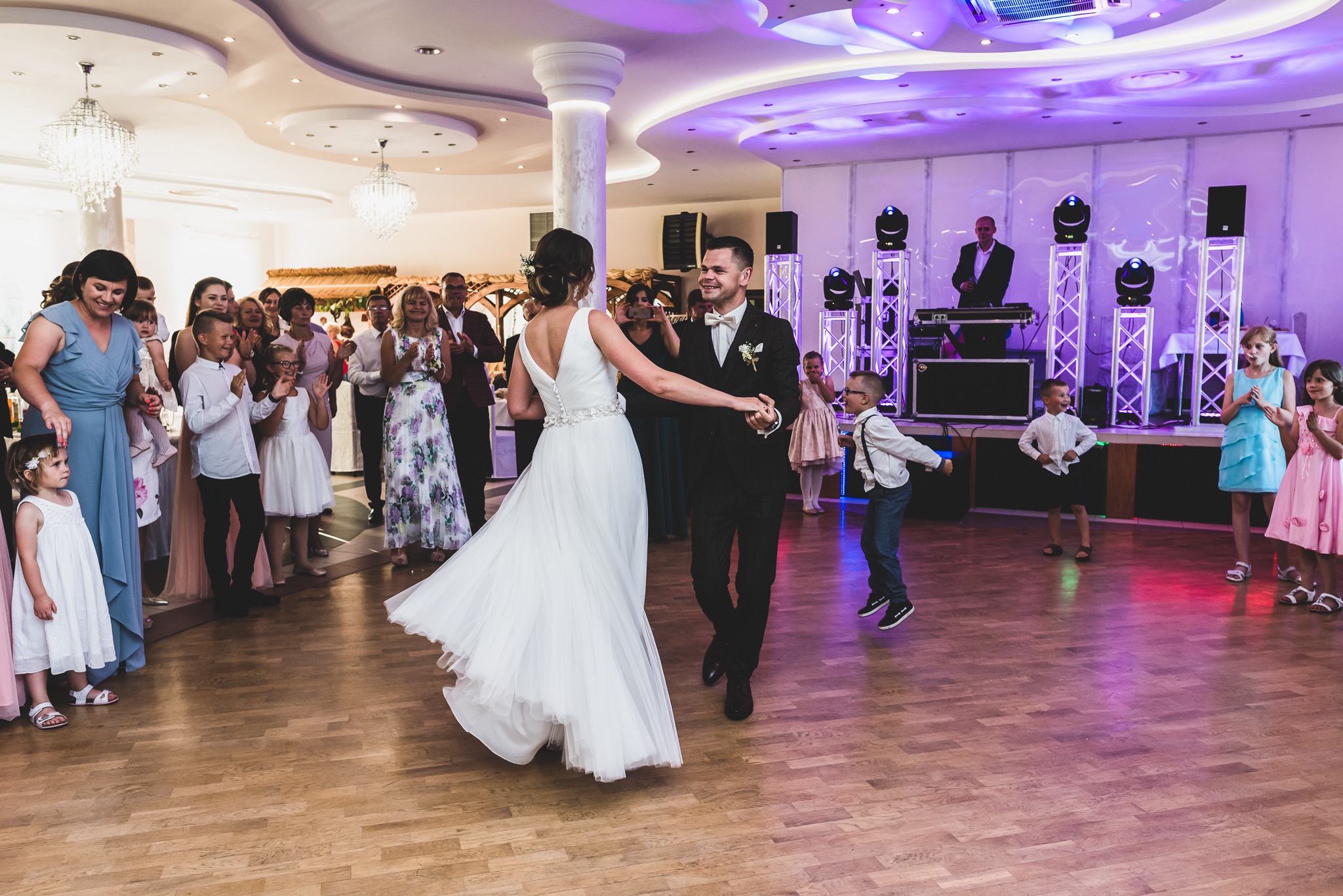 Para młoda tańczy na parkiecie podczas wesela, goście klaszczą, a DJ gra na scenie z oświetleniem.