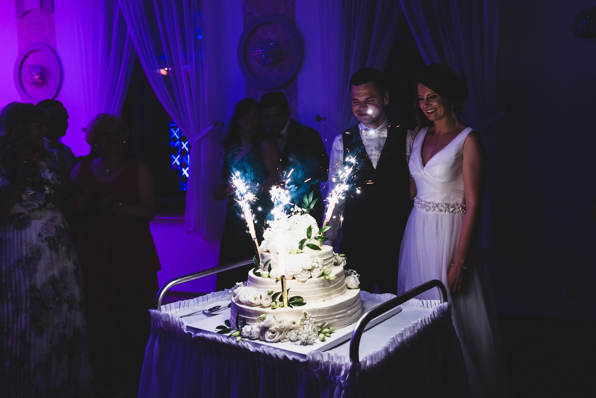 Uśmiechnięta para młoda stoi obok tortu weselnego z fontannami iskier, w tle goście i fioletowe oświetlenie sali.