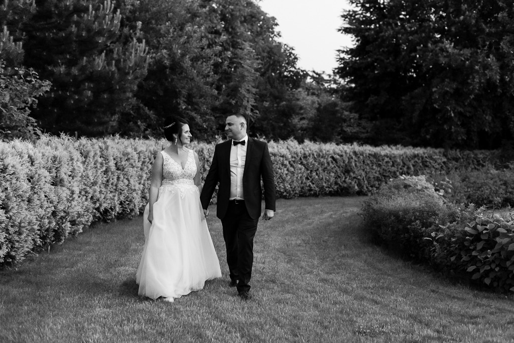 Czarno-białe zdjęcie pary młodej spacerującej po trawniku, panna młoda w sukni z koronkowymi detalami, pan młody w garniturze z muszką, trzymają się za ręce na tle żywopłotu i drzew.