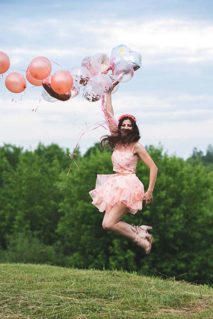 Uśmiechnięta kobieta w różowej sukience i wianku na głowie skacze na trawie, trzymając w ręku balony w odcieniach różu i przezroczyste z konfetti, w tle zielone drzewa i błękitne niebo.