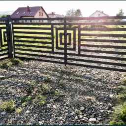 Nowoczesna, metalowa brama dwuskrzydłowa z furtką, pomalowana na czarno, z poziomymi elementami i geometrycznym wzorem na tle żwirowego podjazdu i domów w oddali.