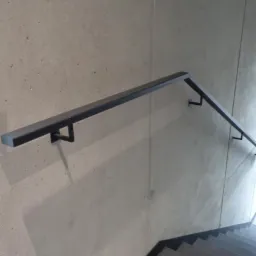 Metalowa poręcz schodowa o kwadratowym przekroju przymocowana do surowej, betonowej ściany wewnętrznej, z widocznym znakiem ewakuacyjnym i fragmentem schodów.