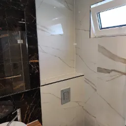 Nowoczesna łazienka z marmurowymi płytkami w odcieniach bieli i czerni, wisząca toaleta z chromowanym przyciskiem, umywalka nablatowa na drewnianym blacie i kabina prysznicowa z czarnymi profilami.