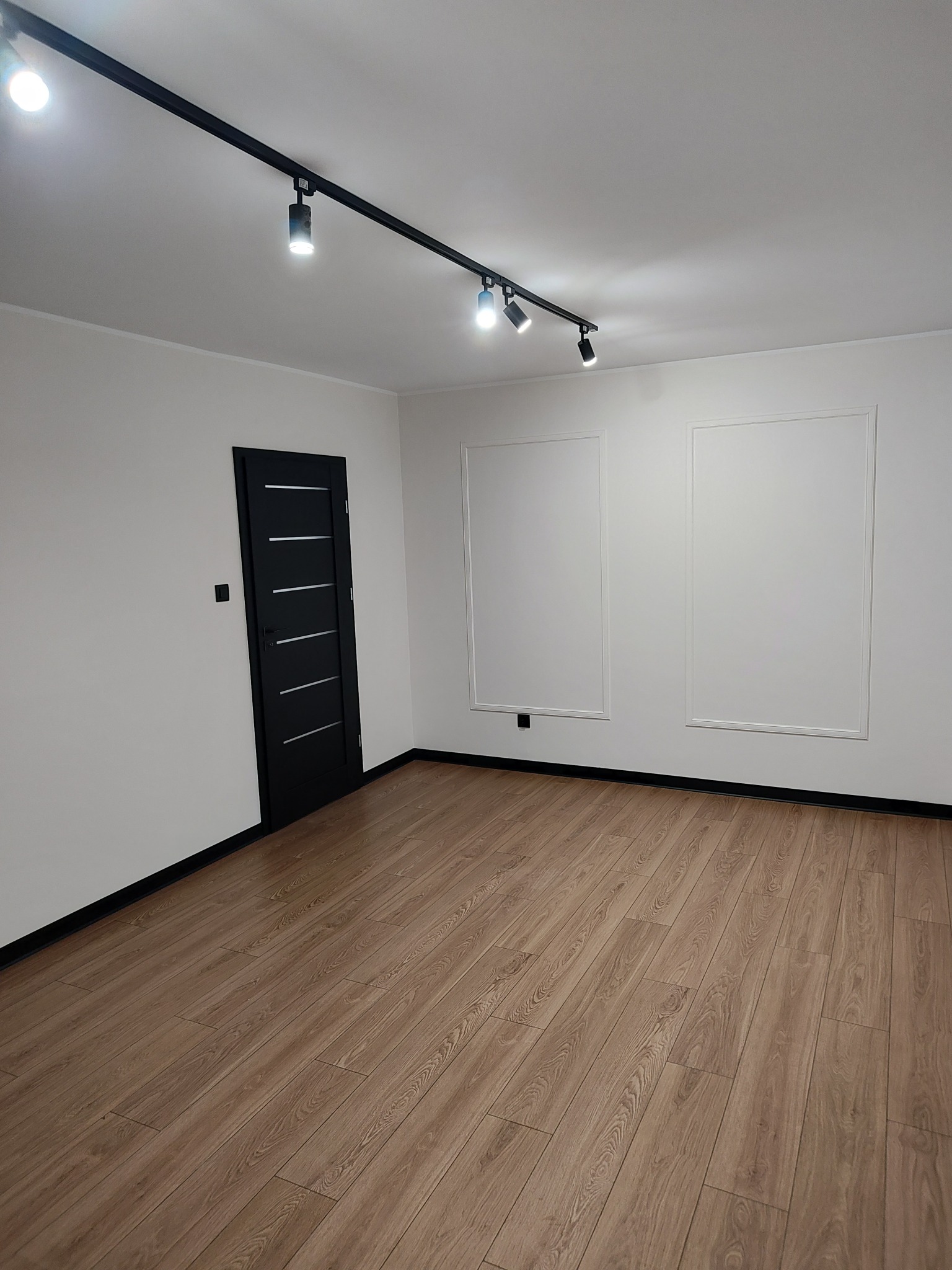 Puste, jasne pomieszczenie po remoncie z panelami podłogowymi w odcieniach brązu, białe ściany z dekoracyjnymi prostokątnymi ramami, czarne listwy przypodłogowe i czarne drzwi z poziomymi srebrnymi...