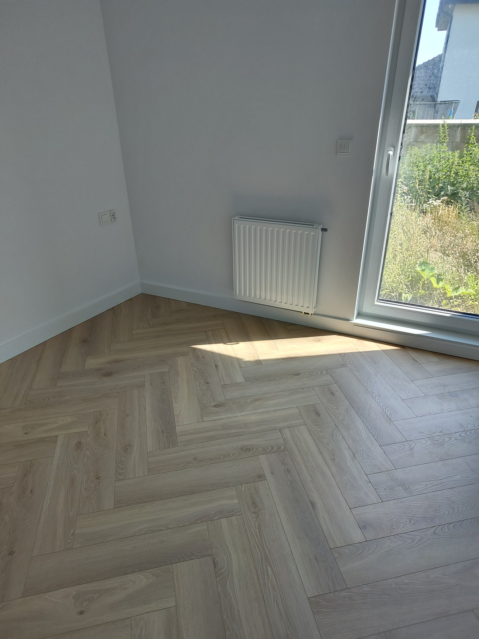 Jasne panele ułożone we wzór jodełki francuskiej w narożnym pokoju z białymi ścianami, grzejnikiem i oknem z widokiem na zieleń na zewnątrz.