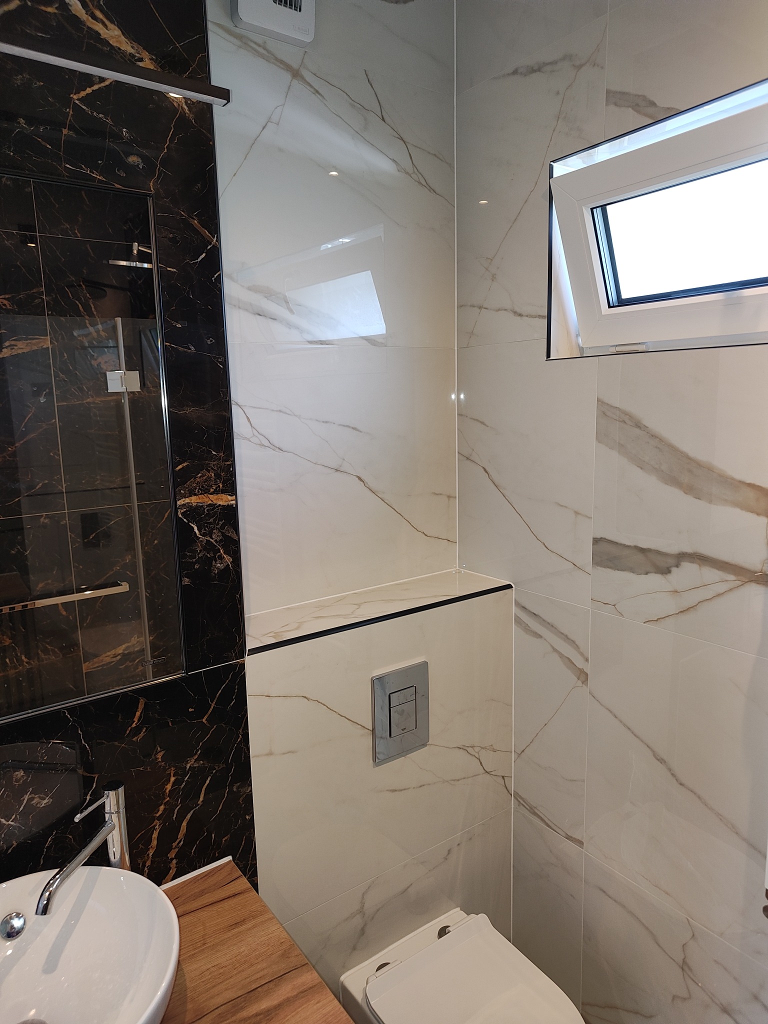 Nowoczesna łazienka z marmurowymi płytkami w odcieniach bieli i czerni, wisząca toaleta z chromowanym przyciskiem, umywalka nablatowa na drewnianym blacie i kabina prysznicowa z czarnymi profilami.