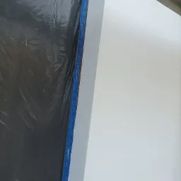 Fragment ściany zabezpieczonej folią malarską i taśmą, sąsiadujący z świeżo pomalowaną, jasną powierzchnią.