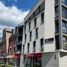 Nowoczesny budynek z lokalami usługowymi na parterze, w tym bank Millennium i salon kosmetyczny BE Beauty, widoczne reklamy i szyldy, chodnikiem przechodzi kobieta.