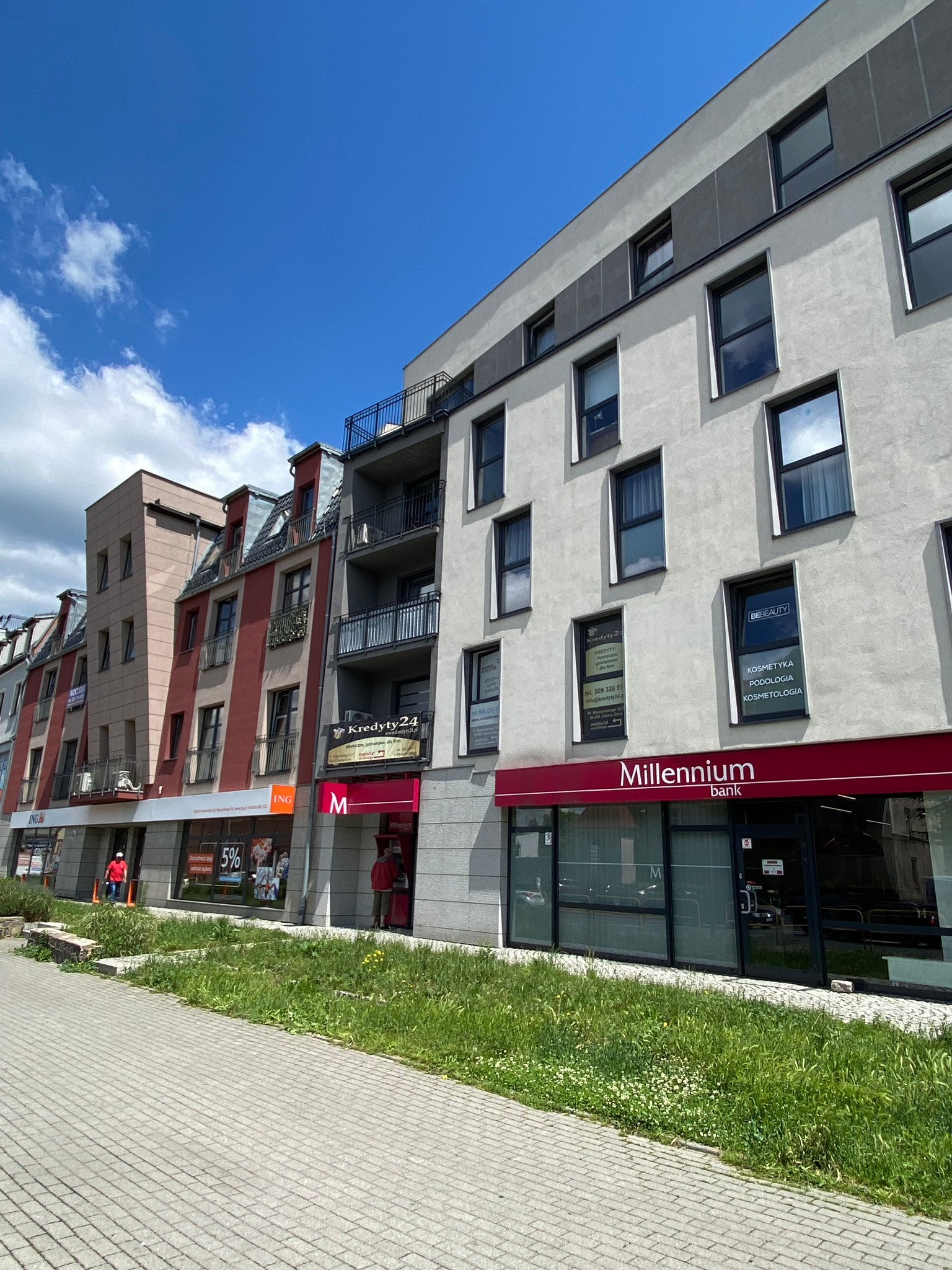 Fasada budynku z lokalami usługowymi, w tym bank Millennium, agencja kredytowa i salon kosmetyczny, widok z poziomu chodnika z kostki brukowej.
